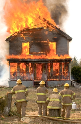 A two-story house is burning, filled with flames that are coming out through the front porch, upstairs windows, and through the roof. Four firemen in gear, with their backs to us, are standing in the foreground, watching the fire.
