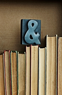 Close up of a section of bookshelf lined with books, spines facing in. The “exposed” pages are facing out and an old letterpress ampersand sits on top of the books, facing the viewer, as if to ask the question, “And….?”
