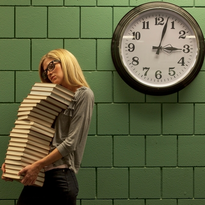 A woman is carrying a huge stack of hardcover books. She is standing in front of an institutional green brick wall on which there is a clock that says 3 o’clock.