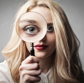 An attractive, long-haired blond woman looks straight at the camera, holding up a magnifying glass in front of her right eye, making that eye appear extremely large within the frame of the magnifying glass.