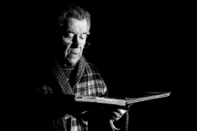 A striking black and white image of an older man with glasses perched on his nose, side lit and wearing a plaid jacket or housecoat, who is holding what looks like an old book and reading.
