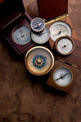 An array of antique compasses, some in wooden casings, arranged haphazardly on a dark brown surface.