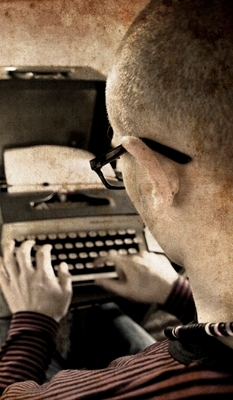 View of a man at his typewriter from over his left shoulder. The man is 30ish, has buzz-cut hair and black-rimmed glasses.