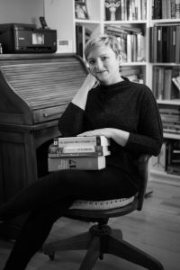 Jahleen Turnbull-Sousa poses in an office chair beside a writing desk with a stack of style guides in her lap. (© Theresa Camastra, Black Dog Studios)